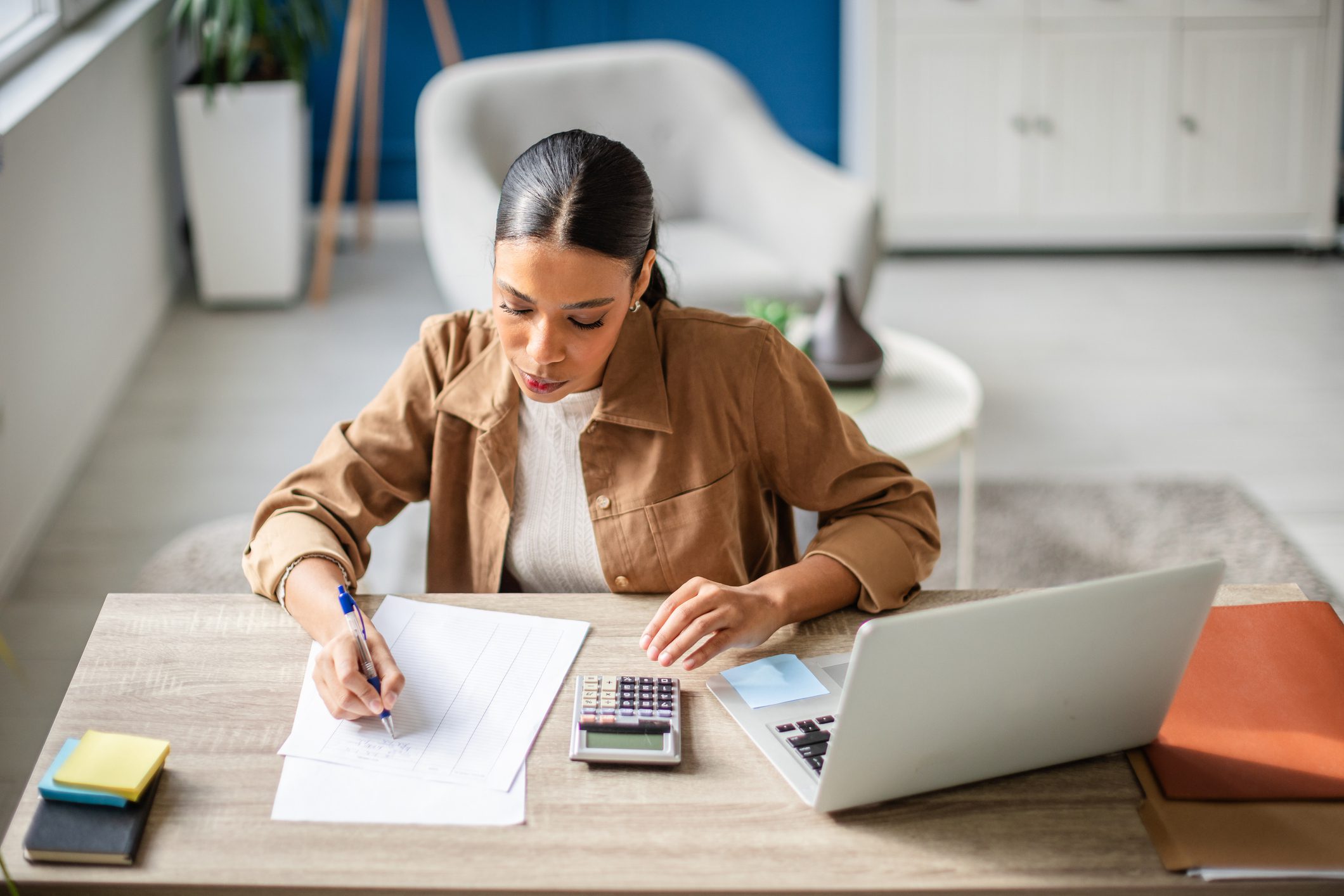 A young hispanic woman is focused on preparing her taxes at a home office. She writes notes, uses a calculator, and organizes her financial documents on the desk.